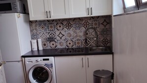A compact kitchen with white cabinets, a washing machine, a stainless steel trash bin, and a patterned backsplash.