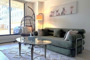 A living room with a green sofa, a glass coffee table, a hanging chair, a lamp, and a wall art piece.