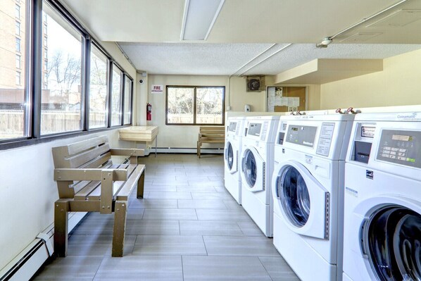 A laundry room with a bench, a fire exit sign, and a row of Coinamat washing machines.