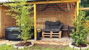 A covered outdoor seating area with a black sofa, a small table, and a chair. There is a bamboo fence and a small tree in the foreground.
