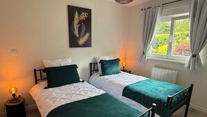 A twin bedroom with two beds, a nightstand, a lamp, a painting, a window with curtains, and a radiator. - At the Secret Garden (Rimsdorf)