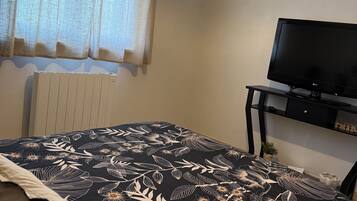 Un dormitorio con cama, televisor sobre un mueble, radiador y ventana con cortinas.