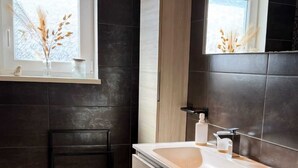 A modern bathroom with dark tiled walls, a wooden vanity, a white sink, and a window with dried plants. - At the Secret Garden (Rimsdorf)