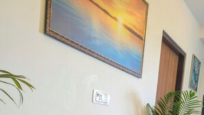 A living room with a beige sofa, a potted plant in the foreground, and a sunset painting on the wall.