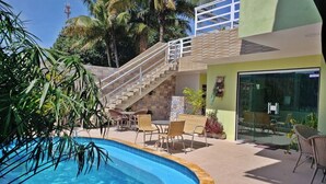 A pool area with a staircase leading to a balcony, surrounded by palm trees and wicker furniture. - 🏡 Roots Inn of Porto! Always ready to welcome you with affection💛📍 (Porto de Galinhas Ipojuca)