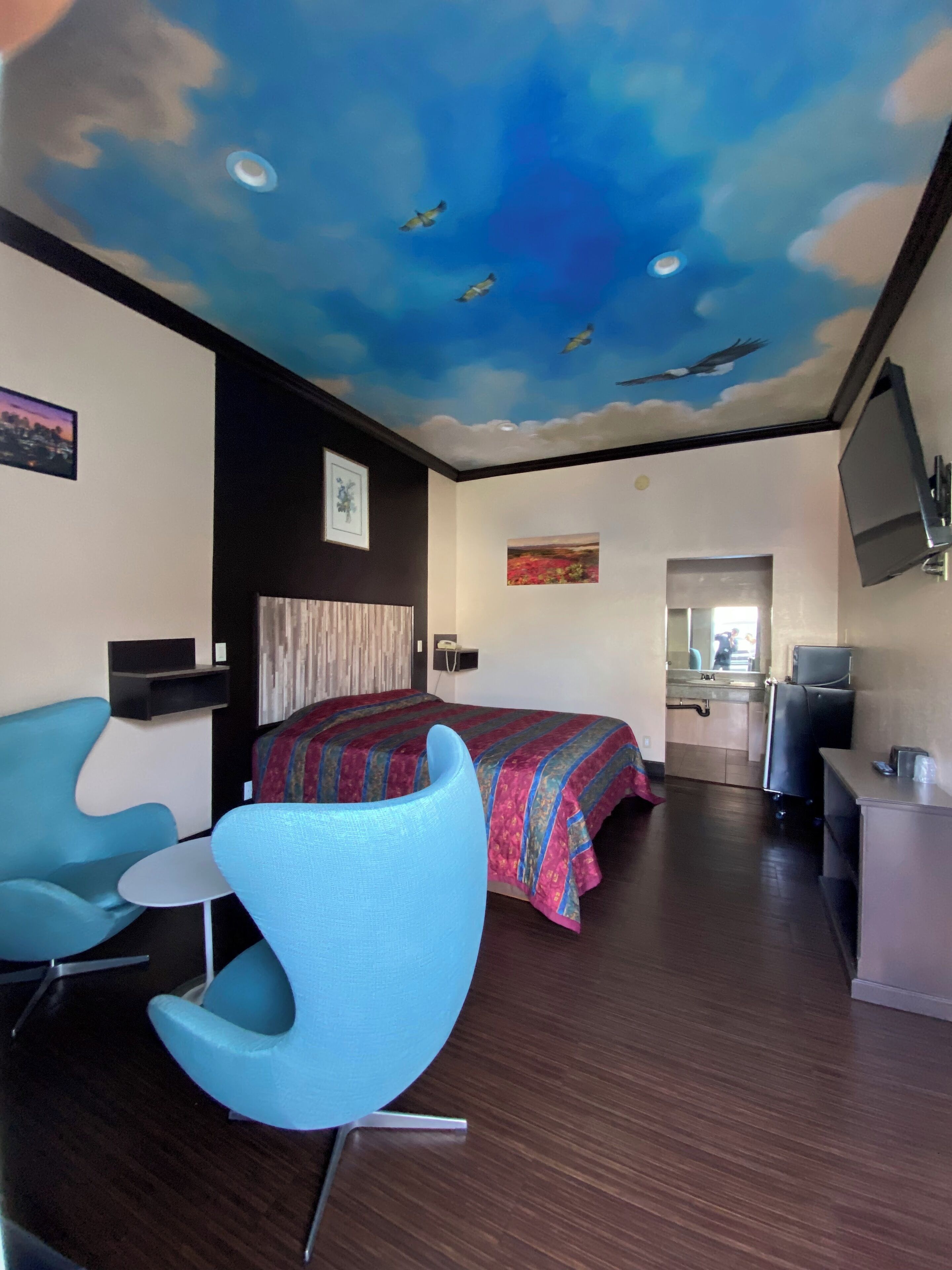 A hotel room with a bed, a blue armchair, a small table, a ceiling mural, a painting, and a TV.