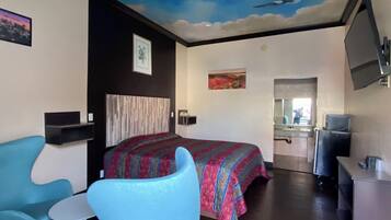 Habitación de hotel con cama, un sillón azul, una mesita, un mural en el techo, un cuadro y un televisor.