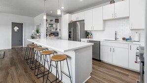 A modern kitchen with white cabinets, a large island with bar stools, stainless steel appliances, and pendant lighting. - Relaxed Island Living Parking, Workspace, AC, WD (Ewa Beach)