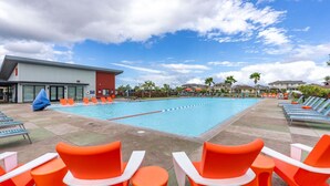 A swimming pool with orange lounge chairs and a covered area, surrounded by buildings and palm trees. - Relaxed Island Living Parking, Workspace, AC, WD (Ewa Beach)