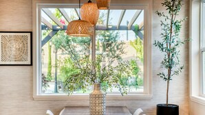 Uma sala de jantar com mesa de vidro, luminárias pendentes de vime, um vaso com folhagem e um vaso de planta.