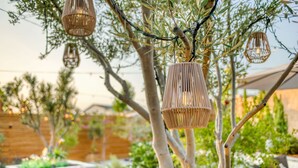 Iluminação suspensa em vime, cerca de madeira, jardim com plantas e um banco.