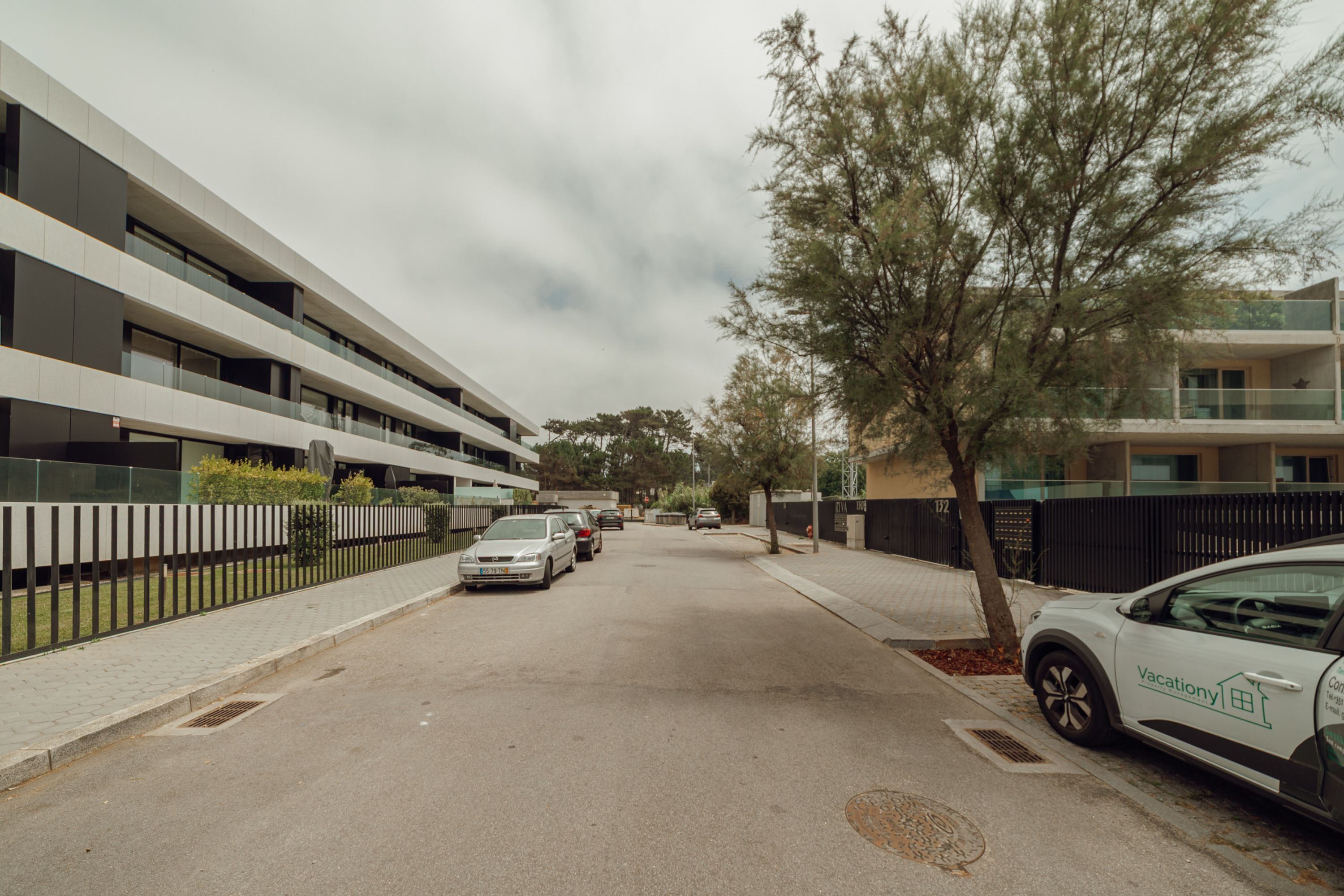 A modern building with multiple floors, a parking area with several cars, and a tree-lined street.