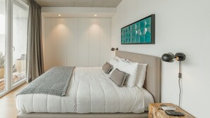 A modern bedroom with a large bed, a bedside table with a lamp, and a view through a sliding door to a balcony.