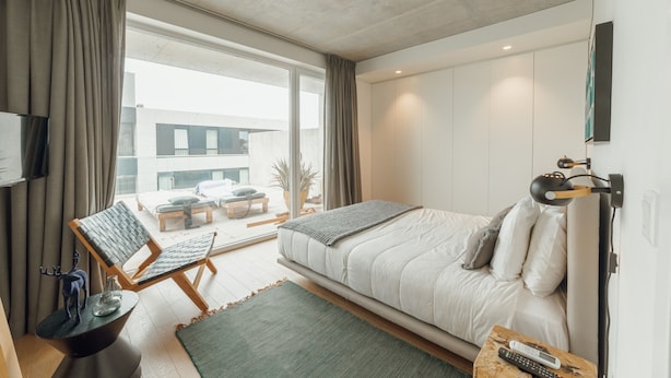 A modern bedroom with a large bed, a seating area with a chair and a small table, and a view of the outside through large windows.