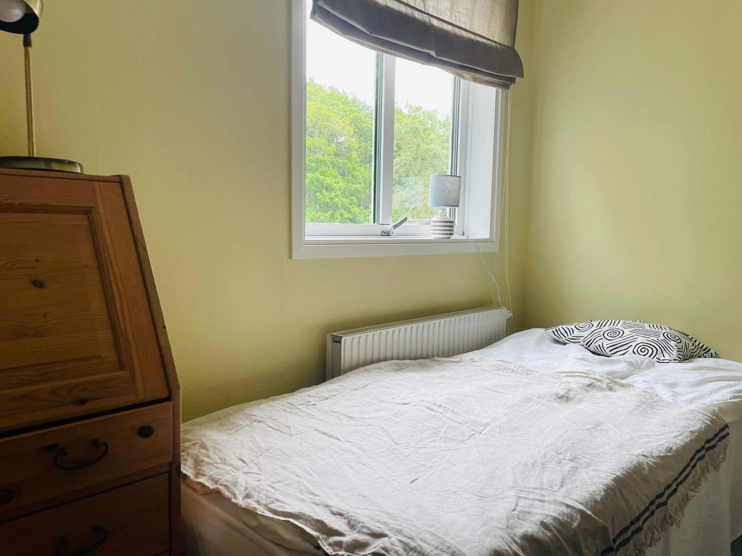 Une chambre avec un lit simple, une table de chevet, une commode, une fenêtre donnant sur un espace verdoyant et un radiateur.