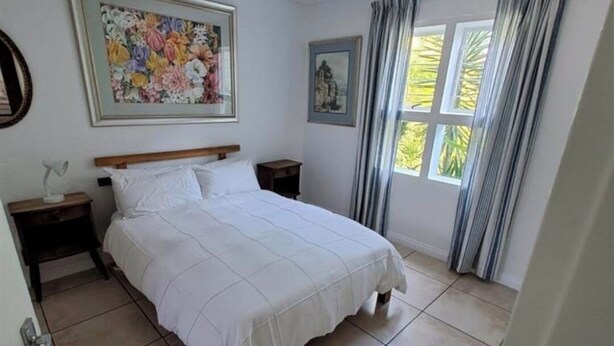 Un dormitorio con una cama grande, dos mesitas de noche, una lámpara en el techo, una ventana con cortinas y un cuadro con flores en la pared.