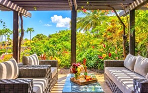 A covered outdoor seating area with wicker furniture, a glass table, and a tropical garden view.