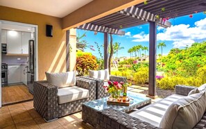 Un patio moderno con mobiliario de mimbre, una mesa de centro de vidrio y vistas a un exuberante jardín con palmeras.