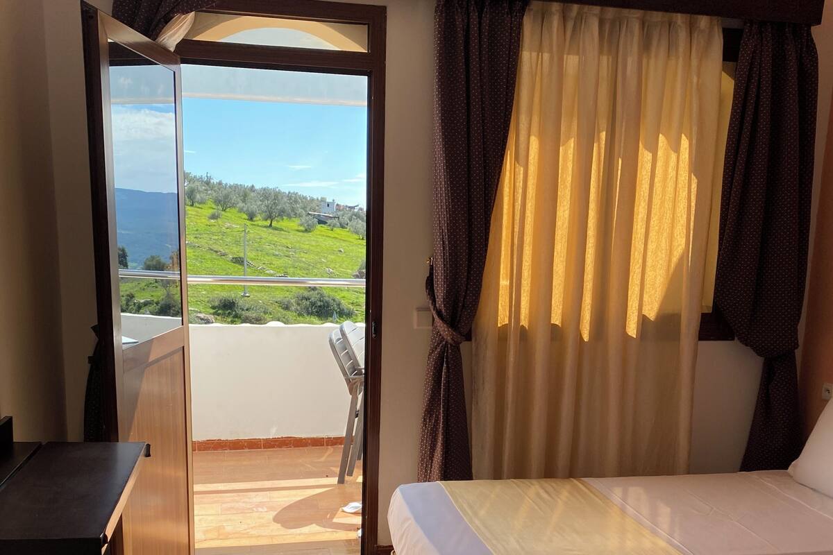 Habitación de hotel con cama, escritorio y vista a un paisaje verde a través de un ventanal.