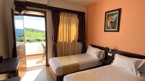 Une chambre d’hôtel avec deux lits, un bureau et une vue sur un paysage verdoyant, visible à travers une grande fenêtre.