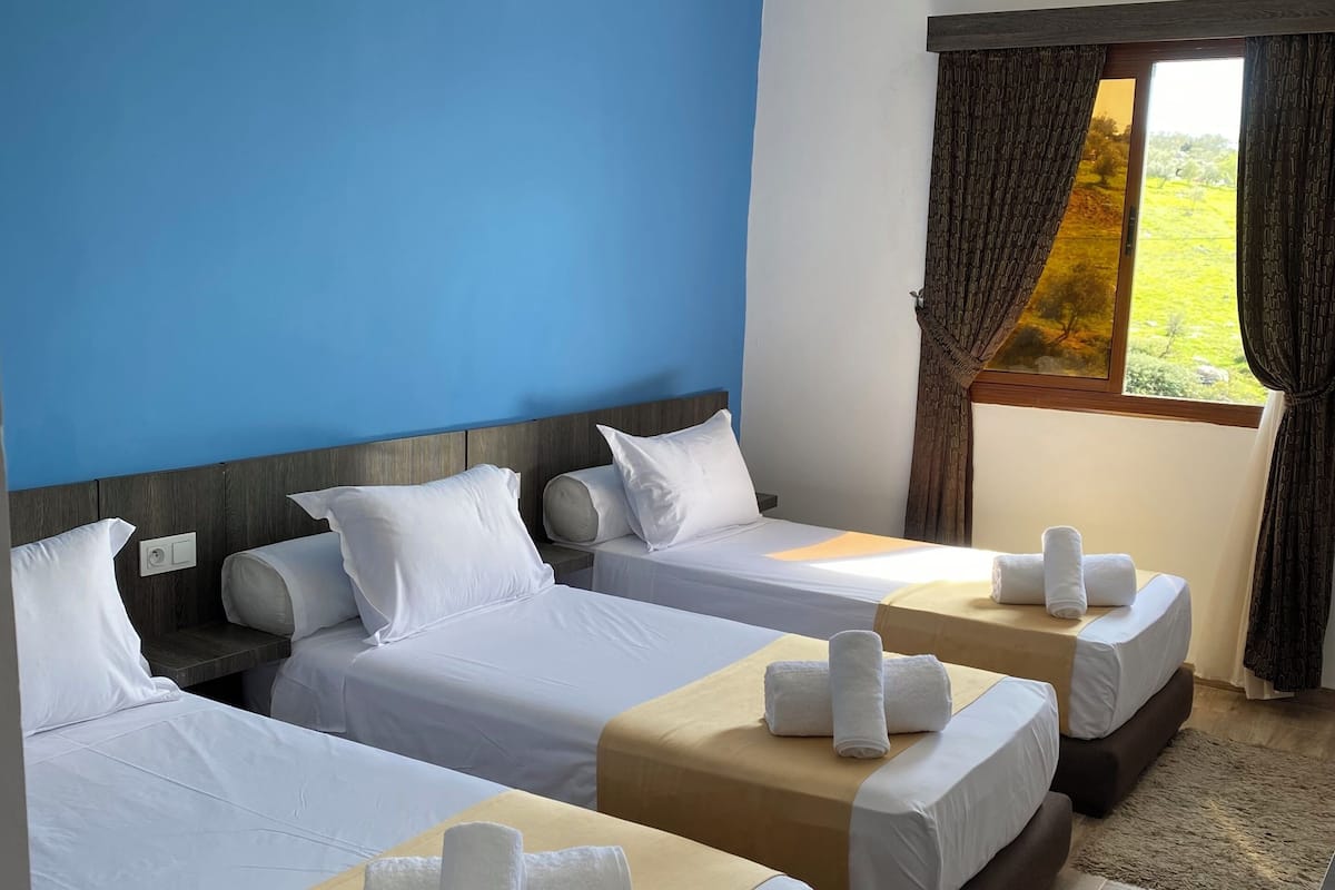 Habitación de hotel con tres camas, un ventanal con cortinas y vista a espacios verdes.
