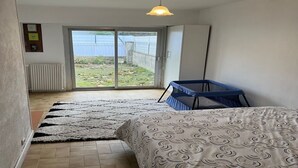 Une chambre avec un lit, un berceau bleu, un tapis à motifs, un radiateur et une vue sur l’extérieur, visible à travers une porte-fenêtre coulissante.