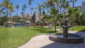 A resort area with a central fountain, palm trees, lounge chairs, and a pool area. - LOCATION LOCATION LOCATION 2 (Cabo San Lucas)