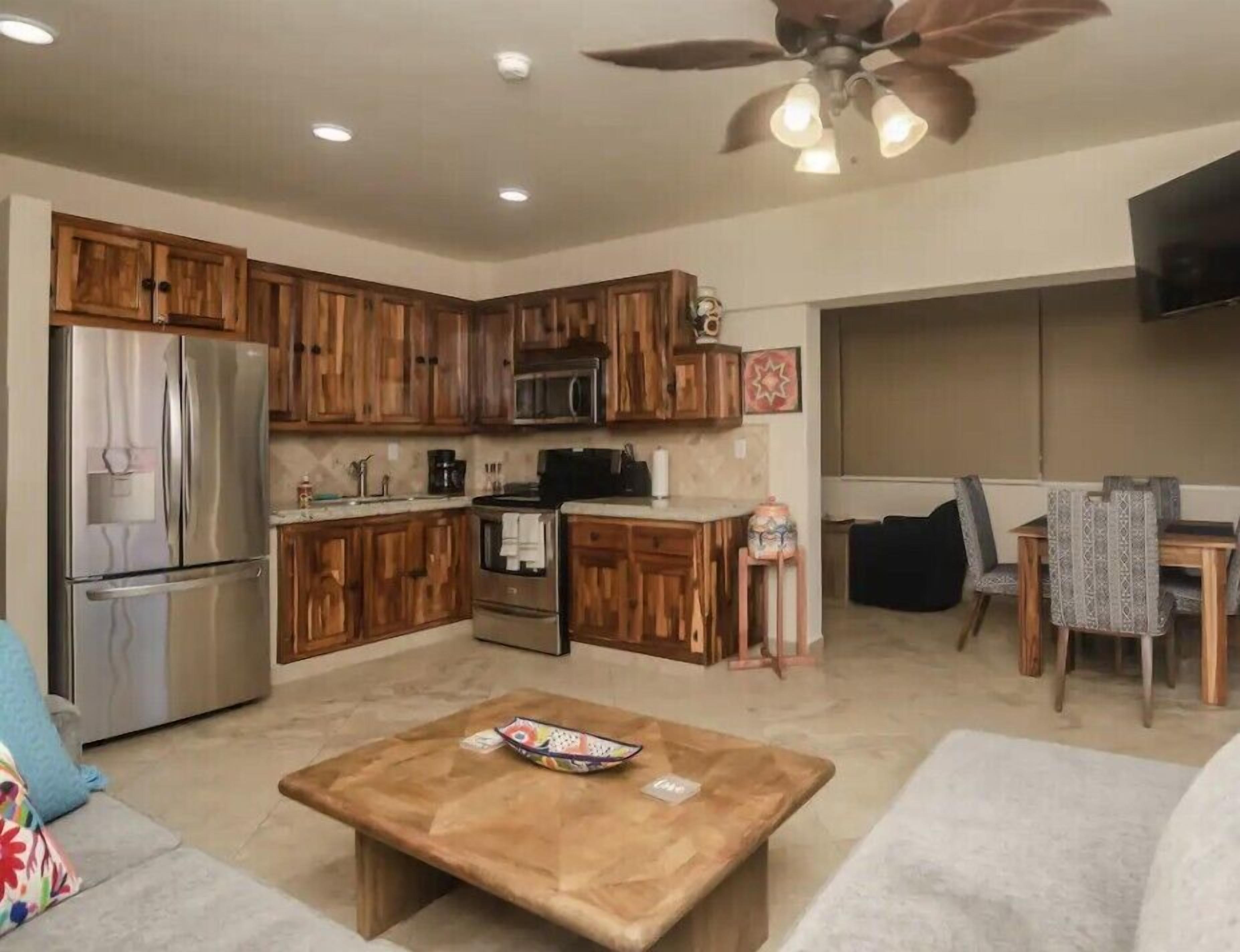 A compact kitchen with wooden cabinets, a stainless steel refrigerator, and a microwave. There is a dining area with a table and chairs.