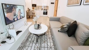 A modern living room with a television, a sofa, a coffee table, and a kitchen in the background. - Bilans Apartments (Accra)