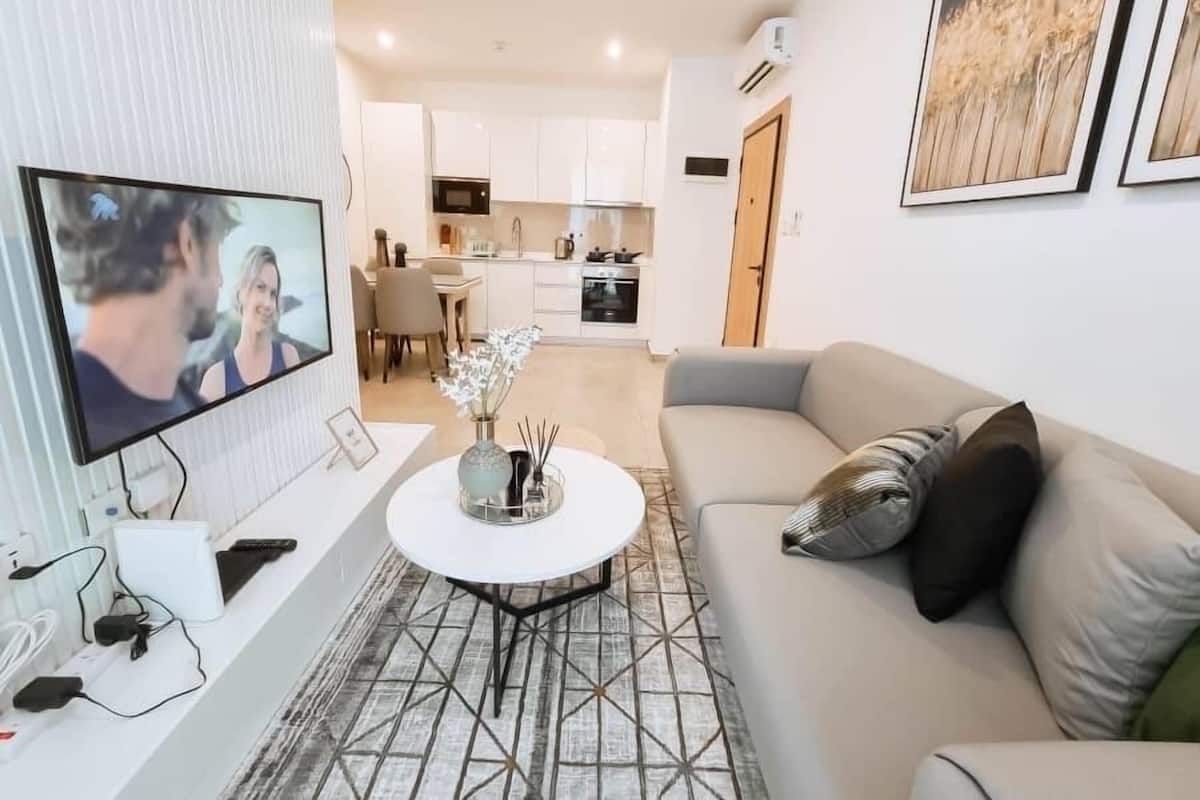 A modern living room with a television, a sofa, a coffee table, and a kitchen in the background.