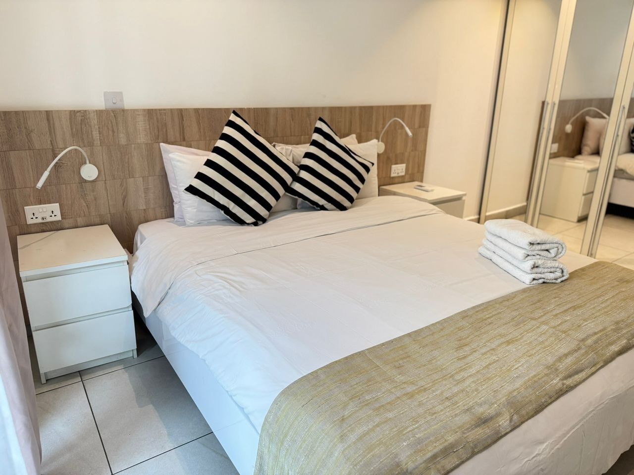A neatly made bed with white bedding, two black and white striped pillows, a bedside table with a lamp, and a mirror on the wall.
