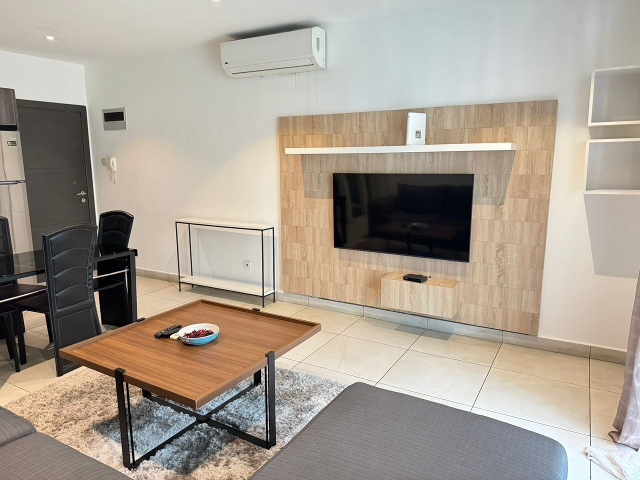 A modern living room with a flat-screen TV mounted on a wooden wall, a dining table with chairs, and a small seating area with a sofa.