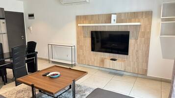 A modern living room with a flat-screen TV mounted on a wooden wall, a dining table with chairs, and a small seating area with a sofa.