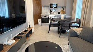 A modern living room with a flat-screen TV, a round coffee table, and a sofa. There is a kitchen area in the background with a dining table and chairs. - Bilans Apartments (Accra)