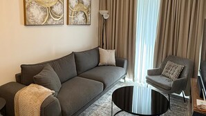 A modern living room with a grey sectional sofa, a round coffee table, and a grey armchair. There are two abstract paintings on the wall and a floor lamp. - Bilans Apartments (Accra)