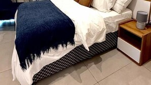 A modern hotel room with a neatly made bed, a wooden headboard, a bedside table with a lamp, and a large window with curtains. - Bilans Apartments (Accra)