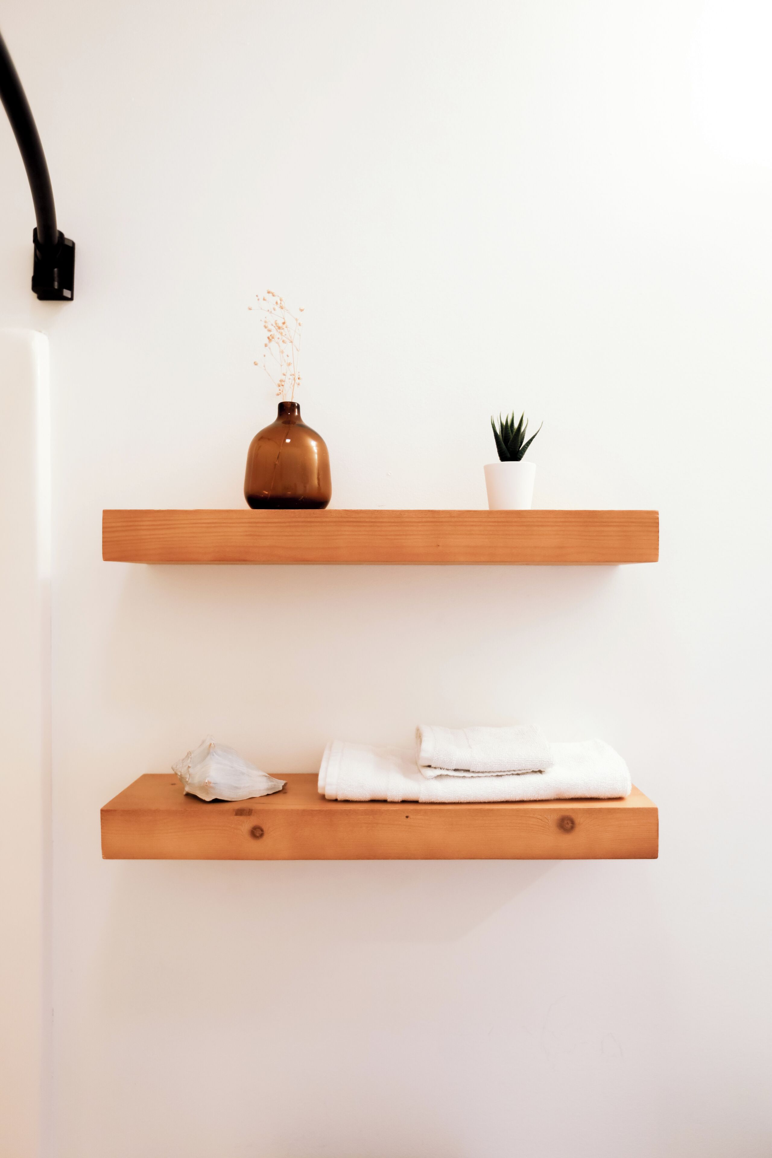 Deux étagères en bois adossées à un mur blanc, l’une avec un vase marron et une petite plante, et l’autre avec des serviettes blanches pliées.
