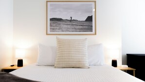 En pænt redt seng med hvidt sengetøj, et indrammet fotografi af en surfer på en bølge, og to sengeborde med lamper på hver side.