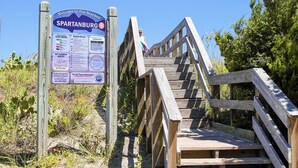 En trætrap fører ned til et strandareal med et skilt der viser strandområdets regler og information.