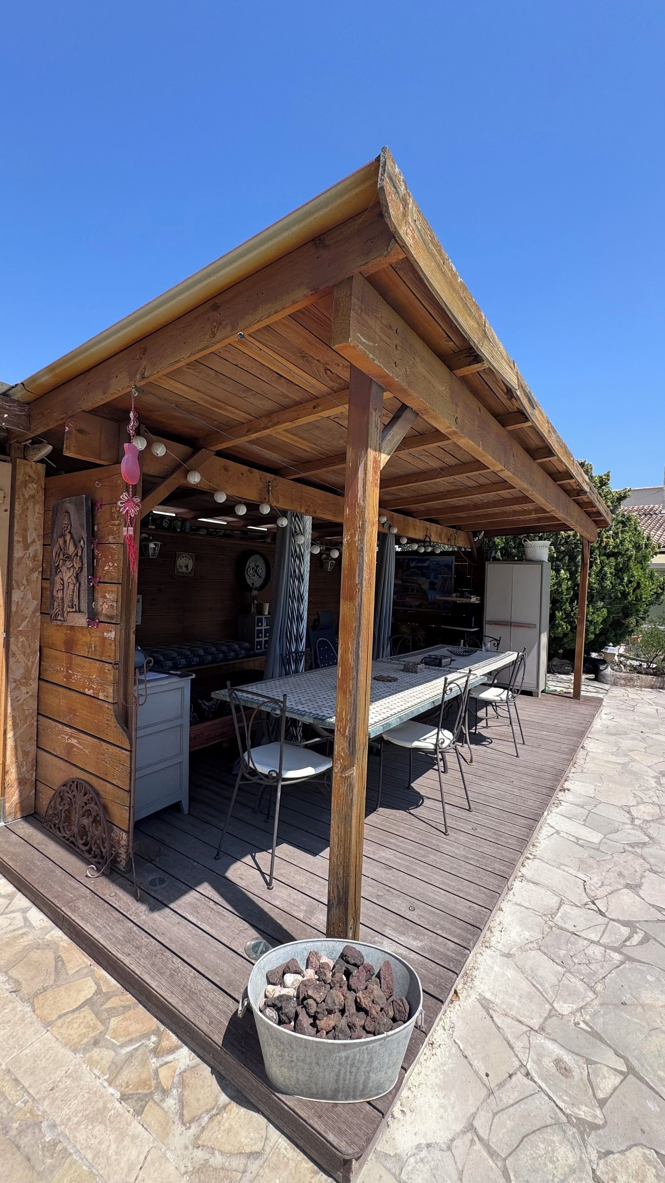 Een overdekt terras met een tafel, stoelen, een emmer kolen en een houten constructie met een rustieke uitstraling.