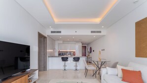 Ein modernes Wohnzimmer mit Fernseher, Sofa und Couchtisch. Es gibt eine Küchenzeile mit Barhocker und einem Esstisch.