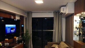 Una sala de estar moderna con un televisor de pantalla plana, un sofá, sillas de mimbre, una mesita y un aparador de vidrio con adornos.
