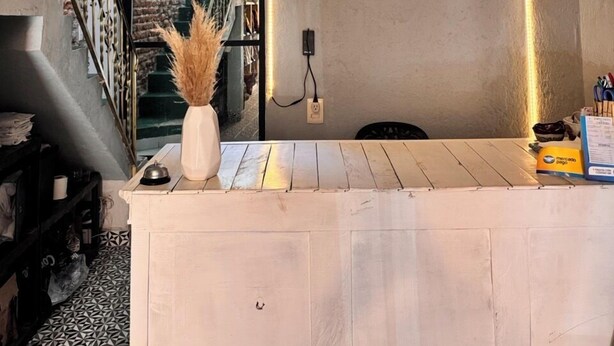 Un comptoir d’accueil d’hôtel avec une enseigne « HÔTEL », un vase contenant des plantes séchées et une vue sur un escalier menant à un mur de pierre.