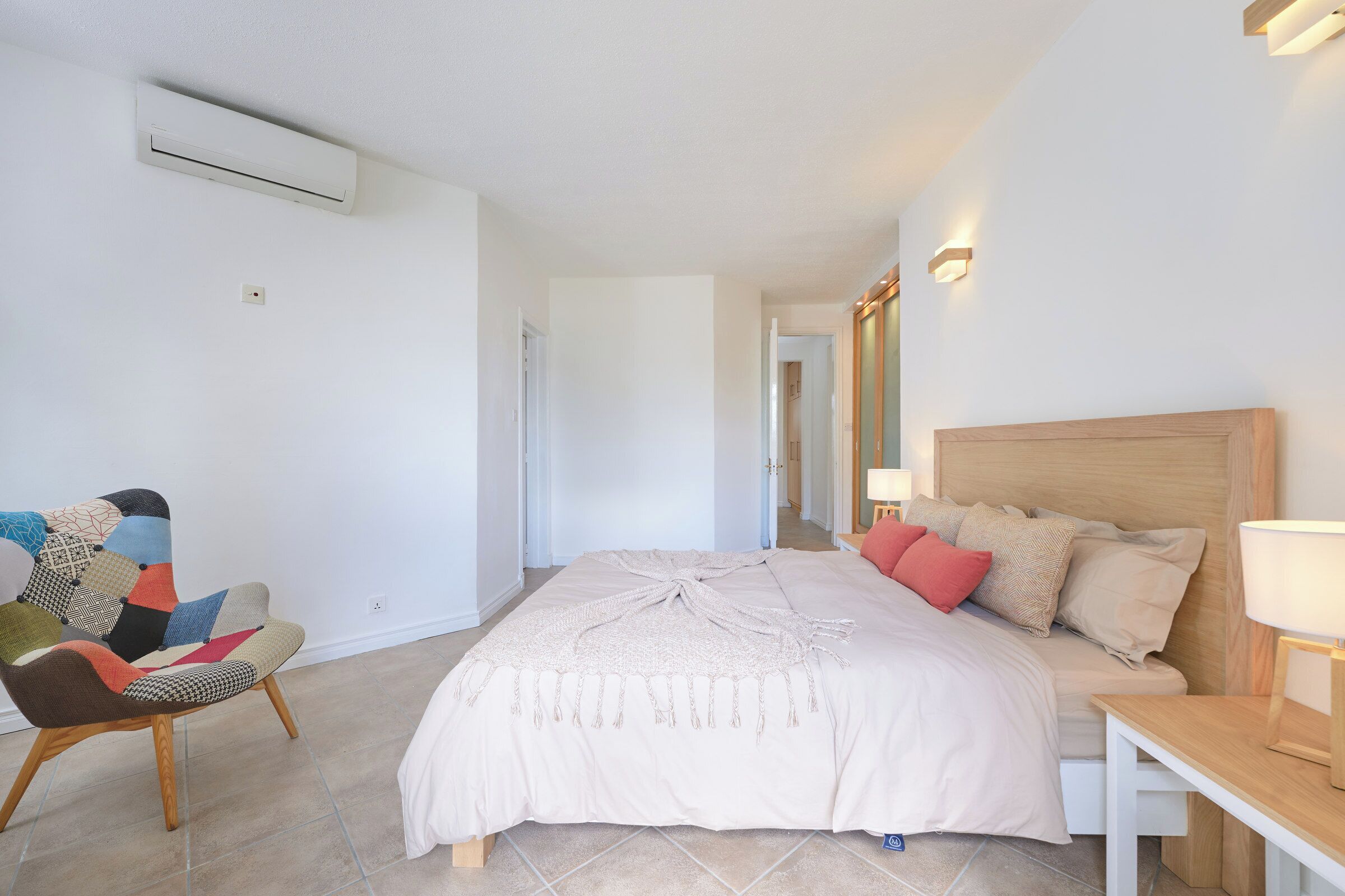 A bedroom with a bed, a chair, a bedside table, and an air conditioning unit.
