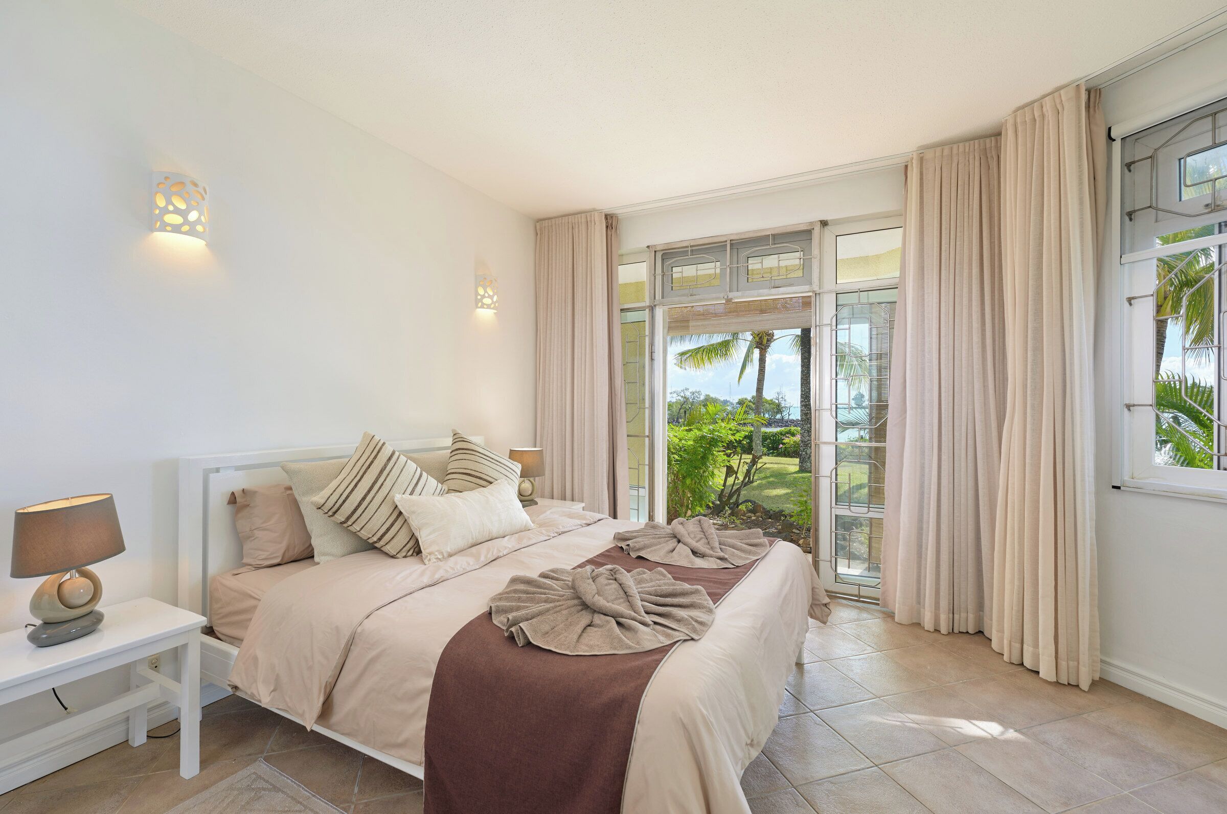 Une chambre avec un grand lit, une table de chevet, une lampe et une vue sur un jardin, accessible par une porte-fenêtre coulissante.