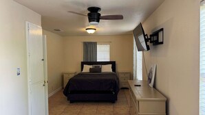 A bedroom with a bed, nightstands, a dresser, a ceiling fan, a wall-mounted TV, and a window with blinds. - Latigo Haven - 7 Mins to Lackland (San Antonio)