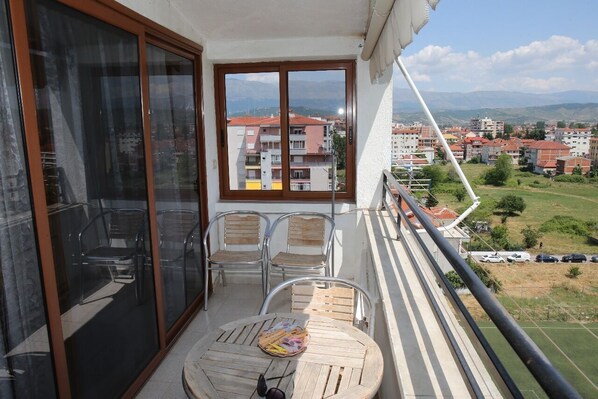 A balcony with a table and chairs, a large window with a view of buildings and mountains, and a railing.