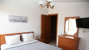 Un dormitori amb un llit, una taula de nit, una làmpada, una petita arca i un mirall, i un quadre penjat a la paret.