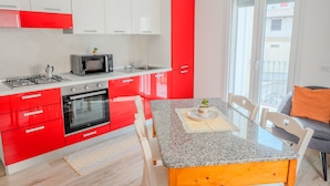 Een moderne keuken met rode en witte kasten, een gasfornuis, een magnetron en een oven. Er staat een eettafel met stoelen en een kleine plant op tafel.