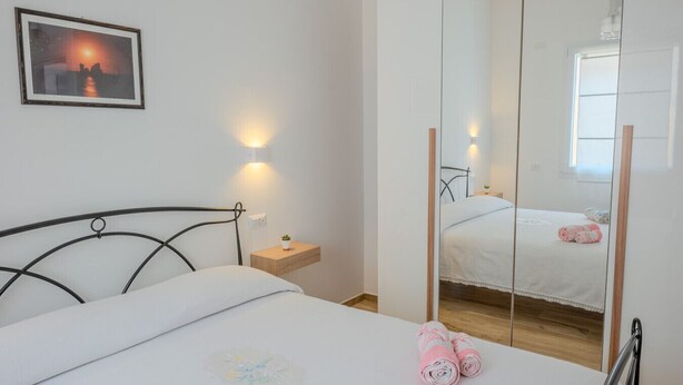 Een slaapkamer met een bed, nachtkastje, een wandlamp en een spiegel die de kamer weerspiegelt.